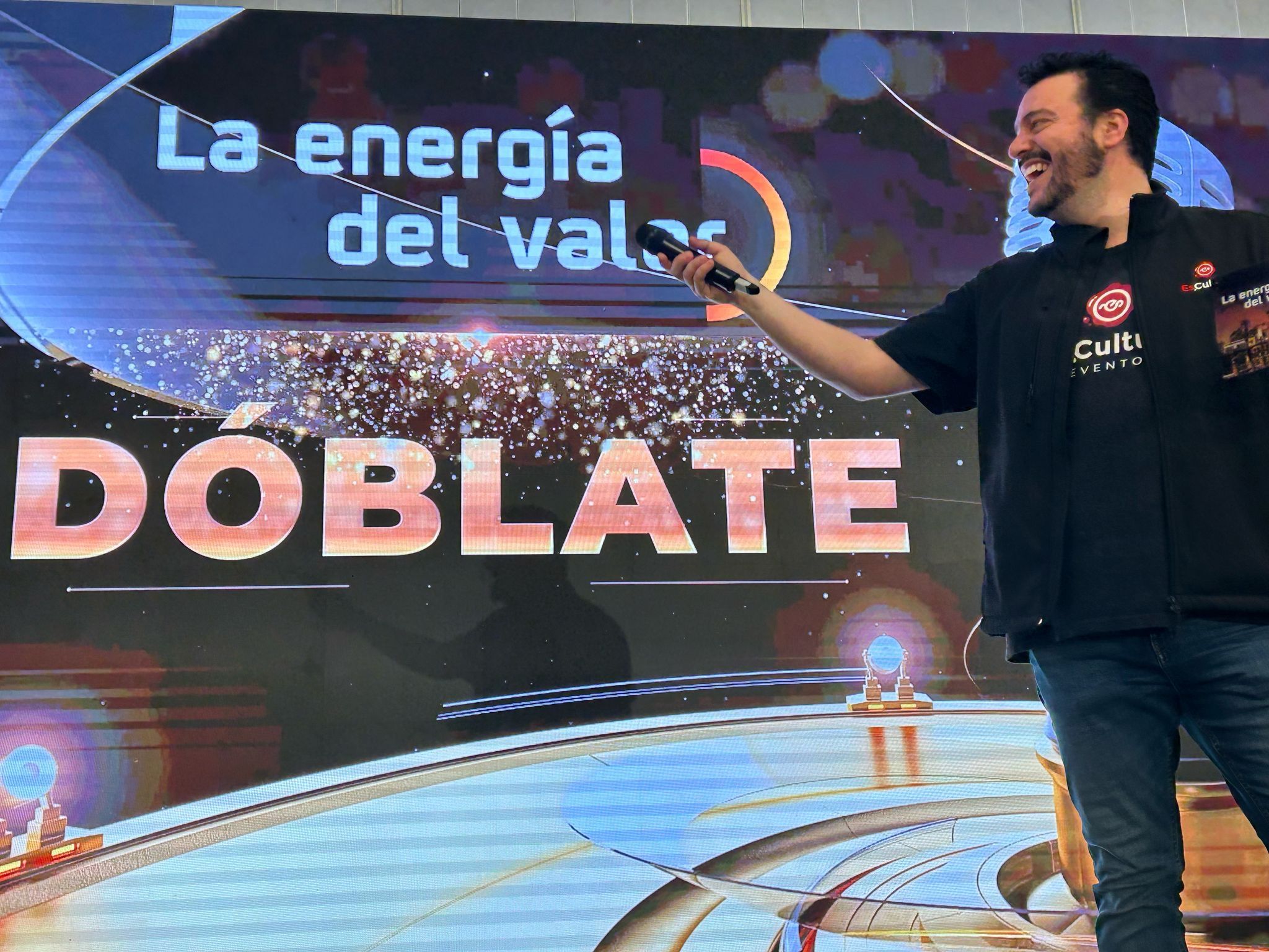 Presentador en el escenario durante la actividad "Dóblate" de Es.Cultura Eventos, con una pantalla que muestra el lema "La energía del valor".